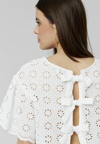 Blouse blanche avec motif de broderie anglaise florale, présentant des manches courtes et une ouverture dans le dos fermée par des nœuds en tissu.