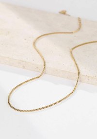 Goldplattierte Kettenhalskette mit einem schlanken, flachen Design. Verfügt über kleine Glieder und eine polierte Oberfläche. Liegt auf einer strukturierten Steinoberfläche.