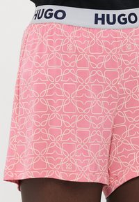 Pink shorts with a geometric cream pattern, featuring a gray elastic waistband with "HUGO" in navy. Made of lightweight fabric.