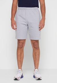 Shorts di lunghezza media grigio chiaro, realizzati in tessuto morbido, con cintura elastica e dettaglio logo laterale. Abbinati a scarpe sportive bianche.