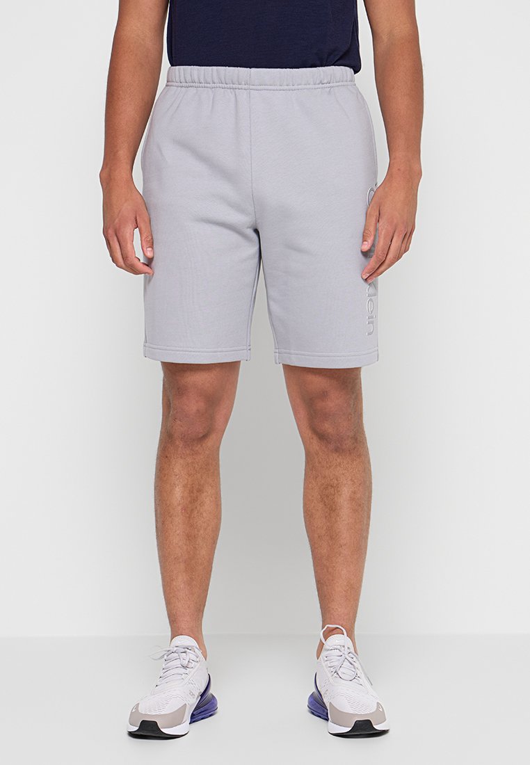 Shorts di lunghezza media grigio chiaro, realizzati in tessuto morbido, con cintura elastica e dettaglio logo laterale. Abbinati a scarpe sportive bianche.