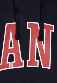 Sweat à capuche bleu marine avec de grandes lettres "MAN" en rouge et blanc. Le tissu est doux, texturé, avec des cordons de serrage au niveau du col.