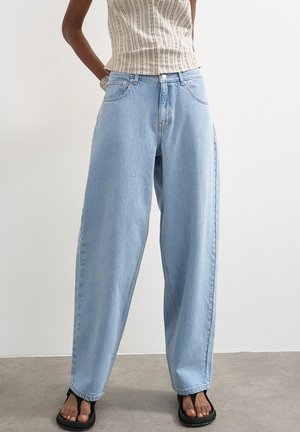 Kvinde iført løstsiddende lyseblå jeans, ærmeløs beige top med striber og sorte sandaler med åben tå, står med hænderne i lommerne.