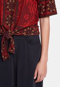 Röd mönstrad blus med korta ärmar och knytbart midjeband, med blommiga och geometriska mönster i gult och svart. Stylad med mörka byxor.