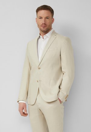 Mann im beigen Anzug und weißem Hemd steht vor einfarbigem hellgrauem Hintergrund, eine Hand in der Tasche, schaut nach vorne.