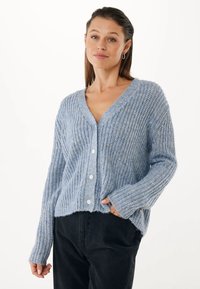 Kobieta w jasnoniebieskim swetrze dzierganym z guzikami, w połączeniu z czarnymi spodniami, stojąca z jedną ręką w kieszeni na białym tle.