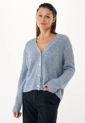 Vrouw in een lichtblauwe gebreide cardigan met knopen, gecombineerd met zwarte broek, staand met één hand in de zak tegen een witte achtergrond.