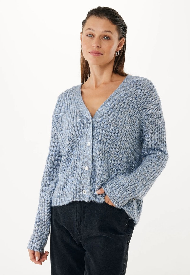 Kobieta w jasnoniebieskim swetrze dzierganym z guzikami, w połączeniu z czarnymi spodniami, stojąca z jedną ręką w kieszeni na białym tle.