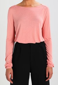 Långärmad topp i ljusrosa tyg med vita prickar, med bred halsringning och samlade ärmar, kombinerad med svarta byxor.