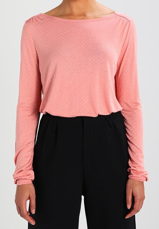 Långärmad topp i ljusrosa tyg med vita prickar, med bred halsringning och samlade ärmar, kombinerad med svarta byxor.