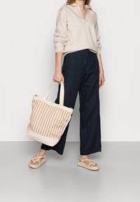 Pull en coton beige avec un col chemise, associé à un pantalon large bleu marine. Tient un sac fourre-tout rayé beige et brun, porte des sandales beiges.