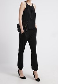 Black sleeveless top with a gathered neckline, paired with loose black trousers and black pointed heels. Accessory is a black clutch with a tassel.