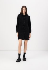 Black corduroy shirt dress with button front, long sleeves, and chest pockets. Model wears black socks and chunky black shoes. Smooth texture.