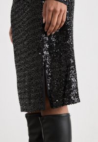 Vestido preto brilhante com superfície texturizada, corte a meio comprimento e fenda lateral. O vestido é combinado com botas pretas até ao joelho.