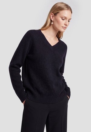 Kvinde med glat blond hår bærer en sort V-hals sweater og sorte bukser, hænderne i lommerne, kigger let nedad og til siden.