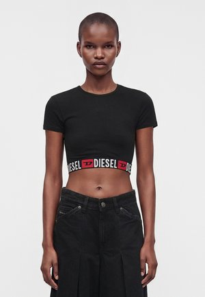 Femme portant un t-shirt court noir avec une bande logo Diesel à la taille et un pantalon noir taille haute, debout devant un fond uni.
