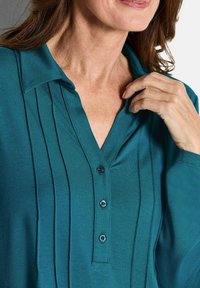 Camicia azzurra con colletto, pieghe verticali e tre bottoni davanti, realizzata in un tessuto morbido con una finitura liscia.
