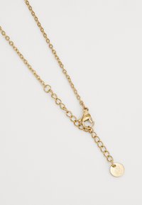 Collier en chaîne dorée featuring des maillons ovales, un fermoir en homard et un pendentif circulaire avec un logo. Texture lisse et finition brillante.
