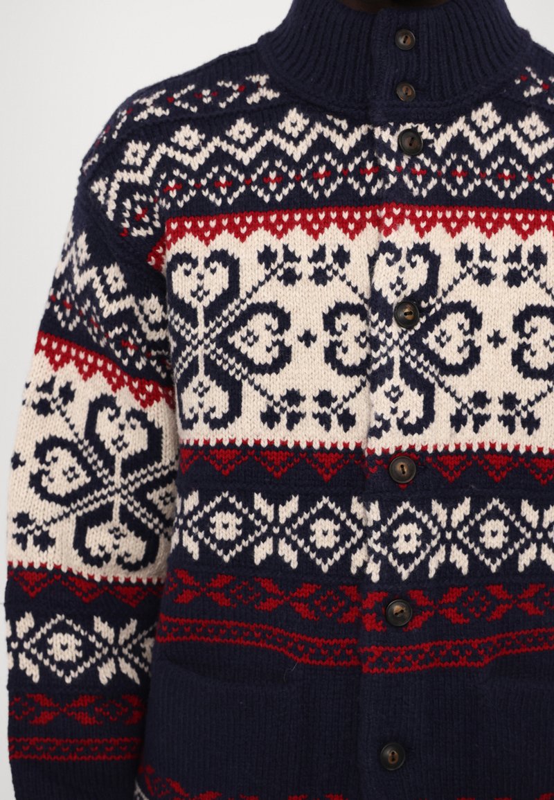 Sweter dziergany z granatowym tłem, wzorami w kolorze kremowym i czerwonym, z płatkami śniegu i sercami, z ribbanym kołnierzem oraz brązowymi guzikami na przodzie.