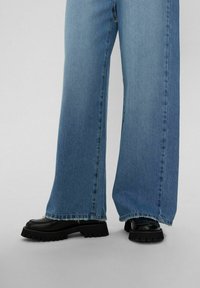 Helle blaue Jeans mit weitem Bein und ausgefranstem Saum, kombiniert mit schwarzen Schuhen mit dicker Sohle. Der Stoff wirkt weich und leicht ausgebleicht.