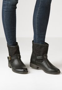 Botas de tobillo negras con parte superior de encaje texturizado, parte inferior de cuero suave y una correa con hebilla dorada. Tacón bajo en bloque y punta redondeada.