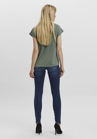 Top scurt cu mânecă scurtă de culoare verde măslin, cu o croială lejeră, asortat cu blugi skinny de culoare albastru închis și tocuri negre, văzut din spate.