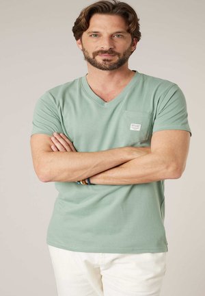 Man met kort bruin haar en baard, gekleed in een lichtgroen V-hals T-shirt en witte broek, met gekruiste armen, tegen een effen achtergrond.