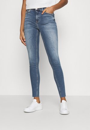 Slim-fit blauwe spijkerbroek met lichte vervaging, rafelige zomen, gedragen met witte sneakers en een wit, ingestopt shirt, hand in de zak.