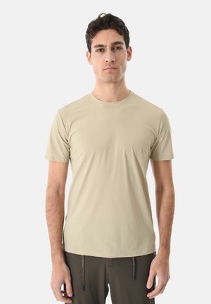 Giovane uomo con capelli scuri corti che indossa una t-shirt beige a maniche corte semplice e pantaloni scuri su sfondo bianco.