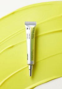 Tube de Retinol Shot Tightening Booster pour une peau ferme sur un fond jaune texturé, avec un emballage argenté et vert et une buse précise.