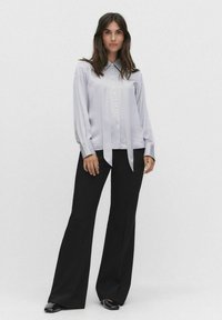 Hellgrau, langärmlige Bluse mit Knopfdetails und einer vorderen Schleife. Kombiniert mit hochtaillierten schwarzen weiten Hosen und schwarzen Schuhen.