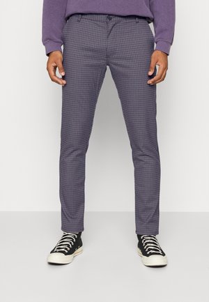 Mężczyzna noszący obcisłe, szaro-czarne spodnie w kratę, w połączeniu z czarno-białymi trampkami, stojący na jednolitym tle.