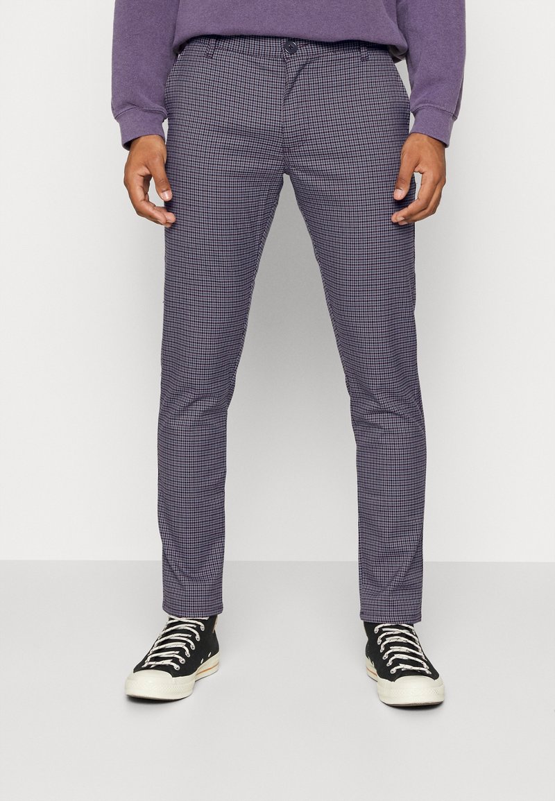 Homme portant un pantalon slim ajusté à carreaux gris et noir, associé à des baskets en toile noires et blanches, debout devant un fond uni.