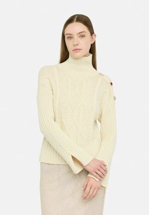 Maglione a dolcevita color crema con texture a coste, motivi a rombi e a treccia, e bottoni decorativi sulla spalla.