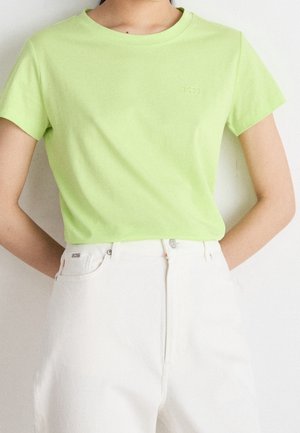 Personne portant une chemise vert citron à manches courtes et un pantalon blanc taille haute, debout les mains dans le dos.