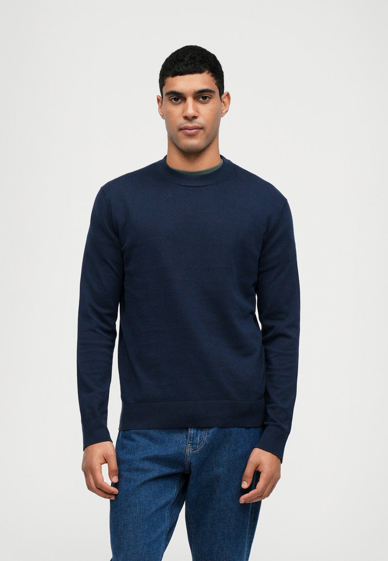 Granatowy sweter crew-neck z dzianiny; posiada długie rękawy i klasyczny krój, sparowany z niebieskimi dżinsami.