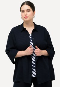 Top nero con bottoni e maniche corte e larghe. Tessuto leggero con una trama sottile, sovrapposto a una camicia a righe blu e bianca.