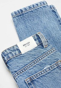 Ljusblå vikta denimjeans med en vit etikett som läser "MNG KIDS EST. 1984" ovanför bakfickan.