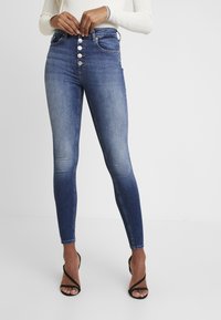 Frau trägt hochgeschnittene, enganliegende blaue Jeans mit Knopfverschluss und schwarze Riemensandaletten, und passt ihre Taille an, während ihr weißes Strickoberteil sichtbar ist.