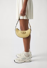 Sac à main en cuir jaune en forme de croissant, avec un fermoir en métal, des bords noirs et des détails de couture. Porté avec une robe blanche.