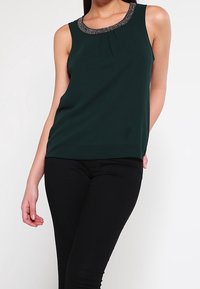 Canotta verde scuro senza maniche con scollatura decorata da perline abbinata a pantaloni neri aderenti, indossata da una persona in piedi su uno sfondo bianco.