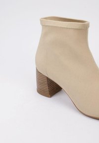 Bota de tobillo de tela beige con un tacón de bloque de madera de tamaño medio, parte superior texturizada y costuras visibles a lo largo del cuello.