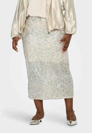 Nő ezüst flitteres midi szoknyában, világos szatén bomber dzsekiben és ezüst alacsony sarkú hegyes orrú cipőben, egyszínű háttér előtt áll.