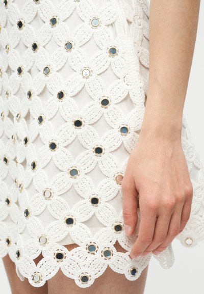 Λευκή φούστα πλεχτή με κυκλικές μαύρες και μεταλλικές διακοσμήσεις, φορεμένη από άτομο με ορατό χέρι που ακουμπά δίπλα στην άκρη της φούστας.