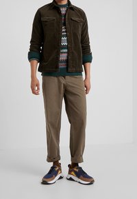 Veste en velours côtelé marron portée sur un pull en maille à motifs, associée à un pantalon beige et des baskets multicolores avec un design marron, bleu et blanc.