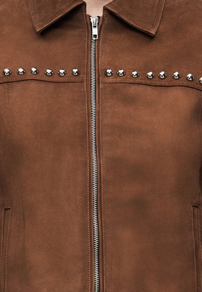 Καφέ σακακι από σουέτ με ασημένια τρουκς στο στήθος και κεντρικό ασημένιο φερμουάρ, με μυτερό γιακά και πλαϊνές τσέπες.