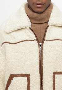 Veste blanche en fausse fourrure avec des accents en cuir brun, dotée d'une fermeture éclair à l'avant et d'un col. Portée par-dessus un col roulé côtelé brun.