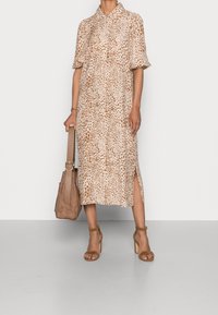 Abito midi beige con motivo leopardato, maniche corte, chiusura a bottoni frontale e fessure laterali, abbinato a una borsa color sabbia e sandali.