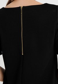 Czarna odzież z okrągłym dekoltem i złotym zamkiem z tyłu, wykonana z gładkiego, fakturowanego materiału.