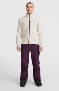 Giacca in pile beige chiaro con zip frontale completa e colletto alto, abbinata a pantaloni cargo viola scuro con tasche laterali. Calzature nere.